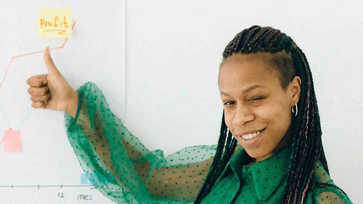 Woman doing the "thumbs up" hand motion in front of a whitebord with drawing of an upward trend graph with a sticker on it with the writing "profit"